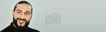 portrait de joyeux homme arabe avec barbe posant en col roulé noir sur fond gris