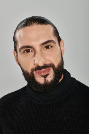 portrait of happy good looking arabic man with beard posing in turtleneck on grey backdrop