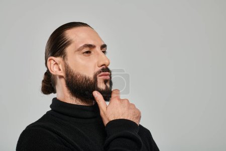 retrato de hombre árabe guapo en cuello alto tocando la barba y posando sobre fondo gris