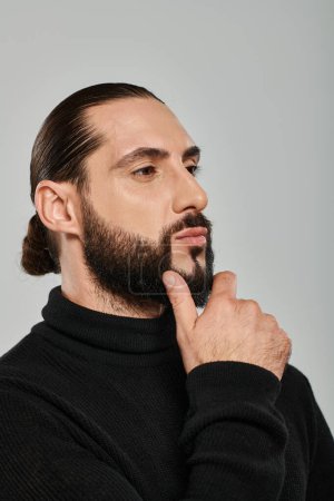 portrait of good looking arabic man in turtleneck touching beard and thinking on grey backdrop