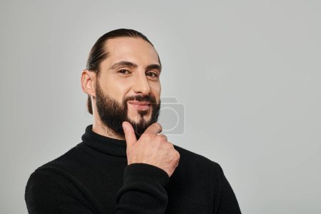 portrait of good looking arabic man in turtleneck touching beard and smiling on grey backdrop