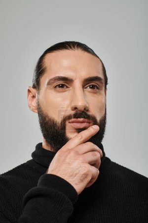 portrait of good looking arabic man in turtleneck touching beard while thinking on grey backdrop