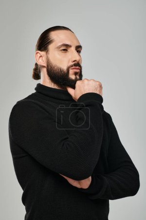 portrait of pensive arabic man in turtleneck touching beard while thinking on grey backdrop