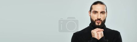portrait d'homme arabe barbu à col roulé noir regardant la caméra sur fond gris, bannière