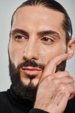 close up shot of confident and bearded arabic man in black turtleneck posing on grey background