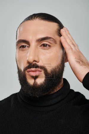 tiro de cerca de hombre árabe barbudo confiado en cuello alto negro ajustando el pelo sobre fondo gris