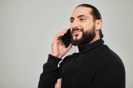 hombre árabe alegre con la barba sonriendo y tener llamada telefónica en el teléfono inteligente sobre fondo gris
