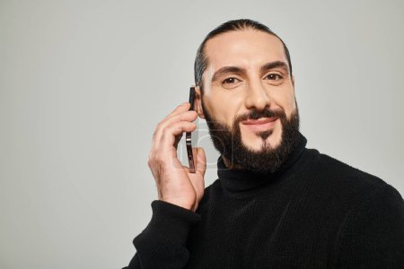 glücklicher arabischer Mann mit Bart, der lächelt und auf grauem Hintergrund mit dem Smartphone telefoniert