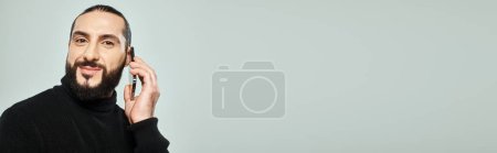 Photo for Happy arabic man with beard smiling and having phone call on smartphone on grey background, banner - Royalty Free Image