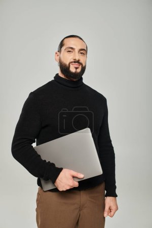 happy and bearded arabic man smiling and standing with laptop on grey background, freelancer
