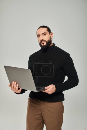 hombre árabe guapo y barbudo de cuello alto negro con portátil sobre fondo gris