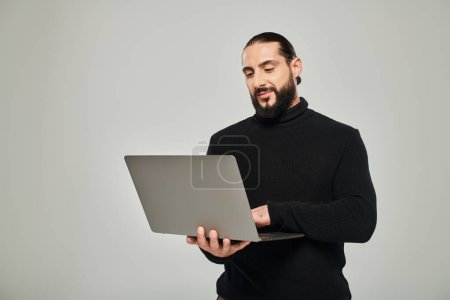 schöner und bärtiger arabischer Mann in schwarzem Rollkragen mit Laptop auf grauem Hintergrund