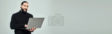Téléchargez les photos : Bel homme arabe barbu au col roulé noir debout avec ordinateur portable sur fond gris, bannière - en image libre de droit