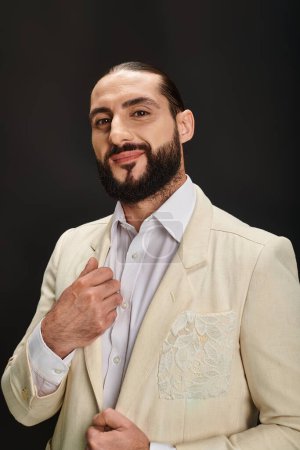 hombre árabe feliz y barbudo en camisa blanca y chaqueta mirando a la cámara sobre fondo negro