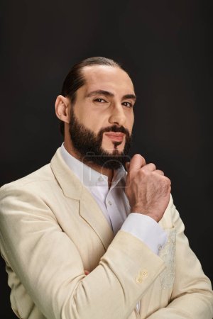 hombre árabe elegante y barbudo en camisa blanca y chaqueta mirando a la cámara sobre fondo negro