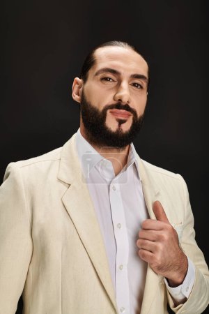 elegant and bearded arabic man in white shirt and blazer looking at camera on black backdrop