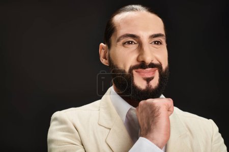 positiver und bärtiger arabischer Mann in weißem Hemd und Blazer, der in die Kamera auf schwarzem Hintergrund blickt