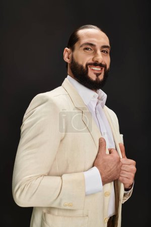 fröhlicher und bärtiger arabischer Mann in weißem Hemd und elegantem Blazer, der auf schwarzem Hintergrund posiert