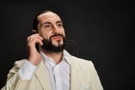 cheerful and bearded arabic man in white shirt and blazer talking on smartphone on black backdrop
