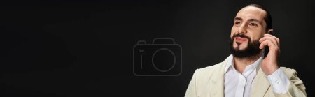 happy bearded arabic man in white shirt and blazer talking on smartphone on black backdrop, banner