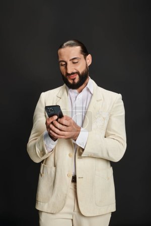 Photo for Cheerful and bearded arabic man in white shirt and blazer texting smartphone on black backdrop - Royalty Free Image
