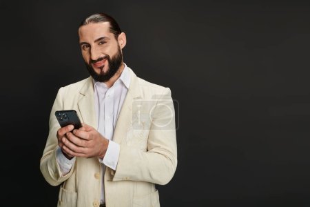fröhlicher und bärtiger arabischer Mann in weißem Hemd und Blazer, der vor schwarzem Hintergrund ein Smartphone textet