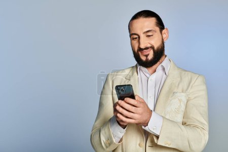 happy bearded arabic man in white formal wear using smartphone on grey background, elegant attire