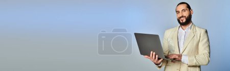 glücklicher bärtiger arabischer Mann in eleganter formaler Kleidung mit Laptop auf grauem Hintergrund, Banner des digitalen Zeitalters