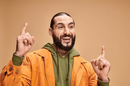 retrato de hombre árabe positivo y barbudo en traje casual apuntando hacia arriba sobre fondo beige