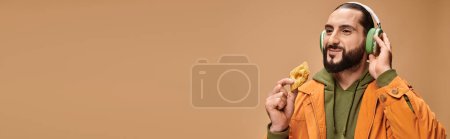 fröhlicher Mann mit Kopfhörern, der Honig-Baklava auf beigem Hintergrund hält, Banner mit mittelöstlichem Dessert