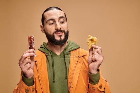 hombre barbudo alegre sonriendo y sosteniendo baklava y cevizli sucuk sobre fondo beige, delicias turcas