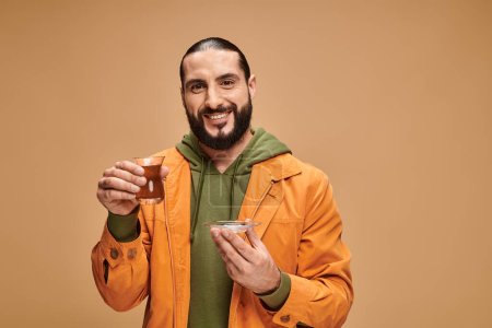 happy bearded man in casual attire holding turkish tea in traditional glass cup on beige backdrop