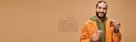 Téléchargez les photos : Heureux homme barbu en tenue décontractée tenant le thé turc dans une tasse en verre traditionnelle sur beige, bannière - en image libre de droit