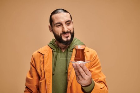 glücklicher bärtiger Mann in lässiger Kleidung, der türkischen Tee in Glasbecher auf beigem Hintergrund betrachtet