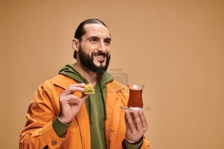 glücklicher bärtiger Mann in lässiger Kleidung mit türkischem Tee in traditioneller Glasschale und Baklava auf Beige