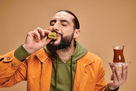 hombre barbudo feliz celebración de té turco en la taza de vidrio tradicional y comer sabroso baklava en beige