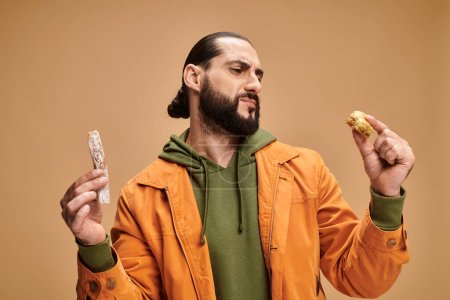 Foto de Confuso hombre barbudo árabe sosteniendo baklava y cevizli sucuk sobre fondo beige, delicias turcas - Imagen libre de derechos