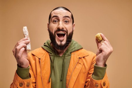 Foto de Hombre barbudo árabe excitado sosteniendo baklava y cevizli sucuk sobre fondo beige, delicias turcas - Imagen libre de derechos