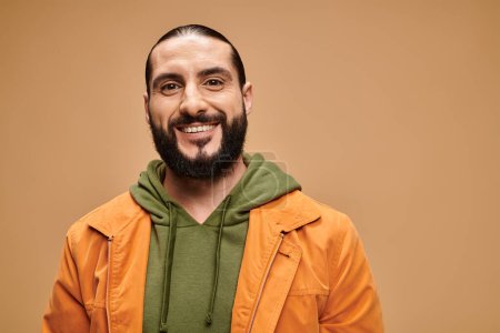hombre árabe feliz con barba de pie en traje casual y mirando a la cámara en el fondo beige
