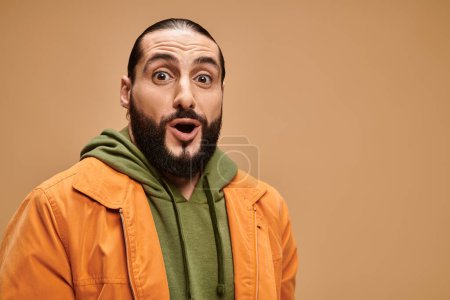 surprised arabic man with beard standing in casual attire and looking at camera on beige backdrop
