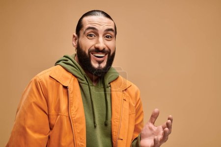 hombre árabe alegre con barba de pie en traje casual y mirando a la cámara en el fondo beige