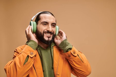 fröhlicher Mann aus dem Mittleren Osten mit Bart, der Musik in drahtlosen Kopfhörern auf beigem Hintergrund hört