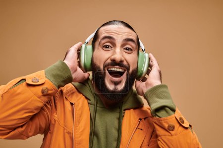 alegre hombre de Oriente Medio con barba escuchando música en auriculares inalámbricos sobre fondo beige