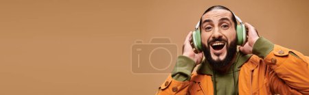 homme du Moyen-Orient gai avec barbe écouter de la musique dans des écouteurs sans fil sur beige, bannière