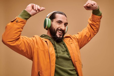 feliz hombre de Oriente Medio con barba escuchando música en auriculares inalámbricos sobre fondo beige