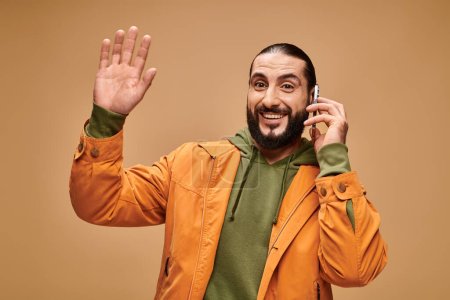 friendly middle eastern man with beard talking on smartphone on beige background, wave hand