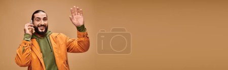 friendly middle eastern man with beard talking on smartphone on beige background, wave hand banner