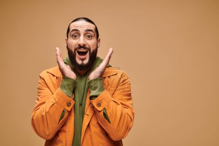 surprised middle eastern man with beard and open mouth gesturing on beige background, wow