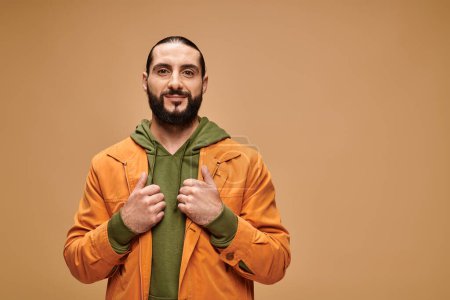 pleased middle eastern man with beard standing in casual attire on beige backdrop, looking at camera