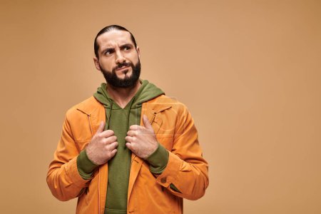 pensive middle eastern man with beard standing in casual attire on beige backdrop, looking away